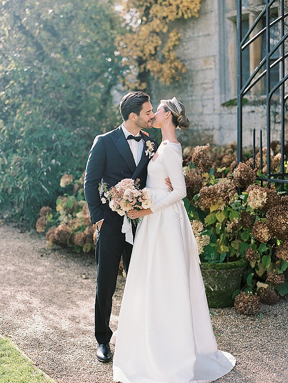 Wedding kiss portrait of bride and groom kissing, bride holding a bouquet in tiara and long-sleeve gown by stone building and greenery