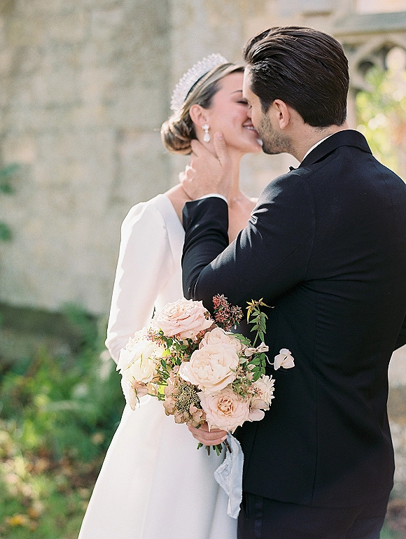 Wedding kiss portrait of bride and groom kissing, groom holding her face, blush rose bouquet and pearl earrings by a stone wall with greenery