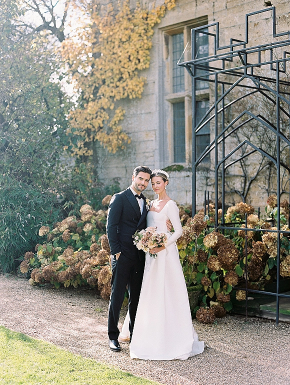 Couple portrait of bride and groom pose, bride holding bouquet in long sleeve dress and tiara beside hydrangeas and stone estate windows