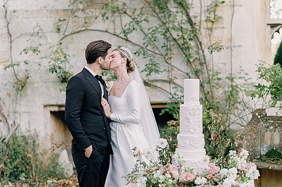 Wedding kiss as bride in veil and tiara and groom in tuxedo stand beside a floral three-tier cake in a vine-covered stone courtyard