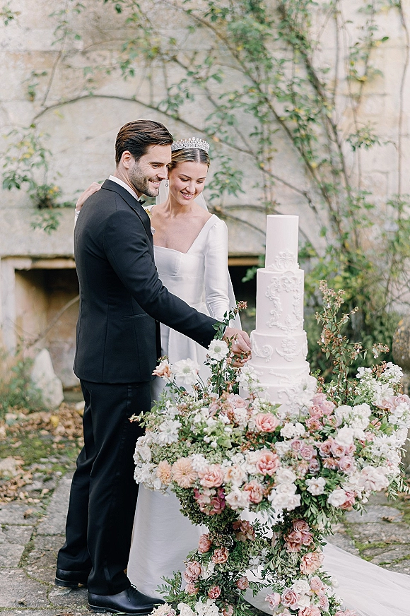 Cake cutting moment as bride and groom hold a knife beside a tiered white wedding cake, bride in tiara and veil in vine-lined courtyard