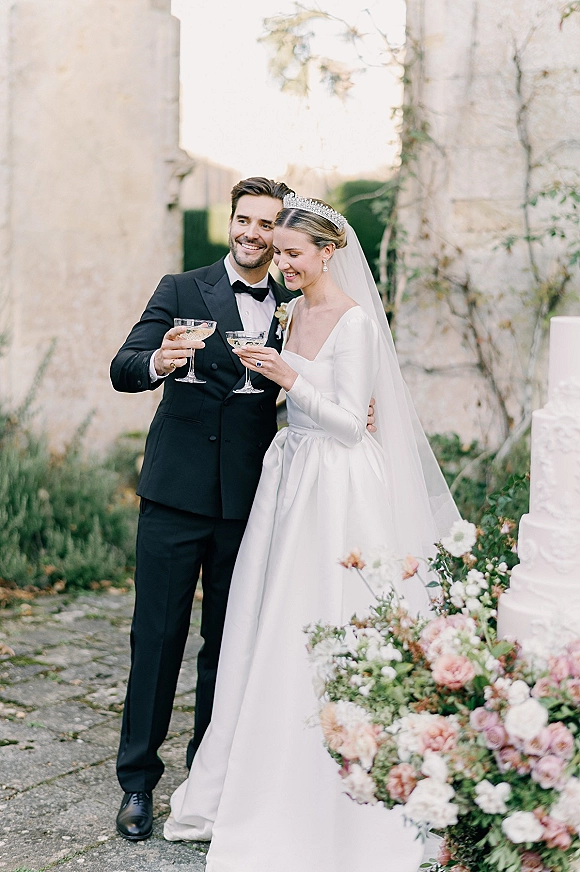 Wedding toast as bride in tiara and veil clinks champagne coupes with groom in black tux beside cake in stone courtyard greenery