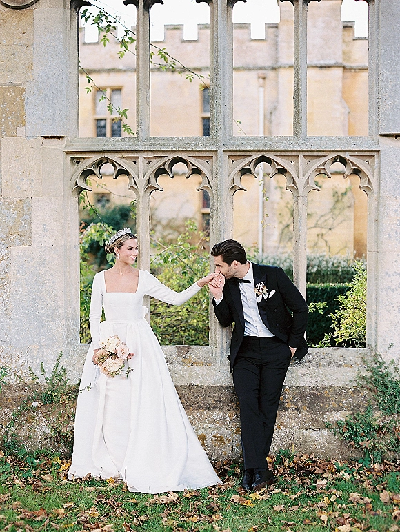 Couple portrait of groom kissing bride’s hand as she holds a blush bouquet, framed by ivy and stone arch windows in a garden lawn