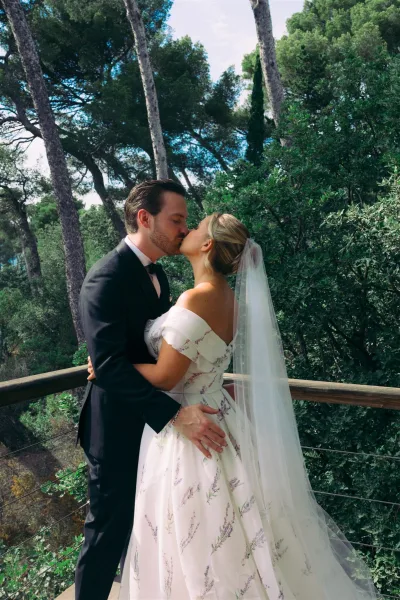 Wedding kiss portrait of a bride with a long tulle veil and her groom embracing, an outdoor wedding portrait by a wood railing amid trees.