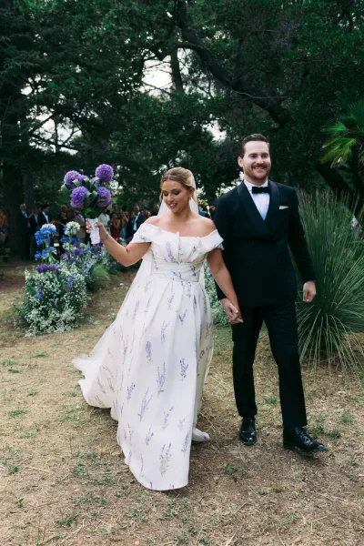 Ceremony recessional newlywed exit with the couple walking hand in hand, bride in a floral print gown, along a garden dirt path under trees.