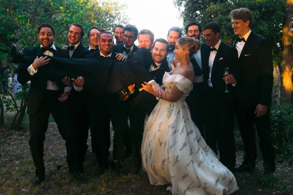 Groomsmen photo with the groom carried by groomsmen, a groomsman holding a champagne flute, against garden trees in warm sunset light.