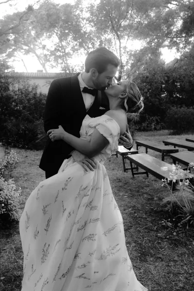 Wedding kiss portrait with a graceful dip kiss wedding pose, groom in a bow tie dipping the bride beneath garden trees beside wooden benches.