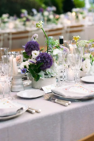 Reception tablescape with purple and white centerpieces and taper candles, set outdoors amid lush garden greenery.