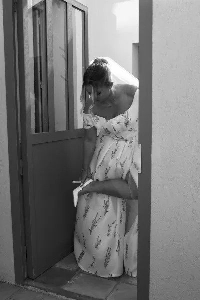 Bride putting on shoes in a quiet getting ready moment, slipping into white heels in a stucco-walled doorway with tile floor.