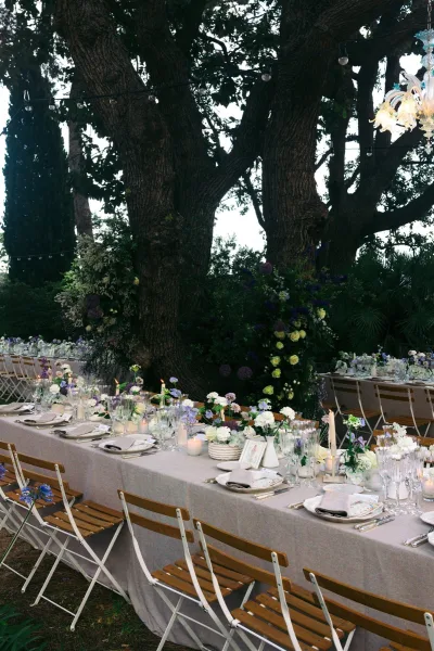 Reception tablescape on an outdoor wedding reception table with taper candles, set beneath large trees and garden greenery, elegant and inviting.