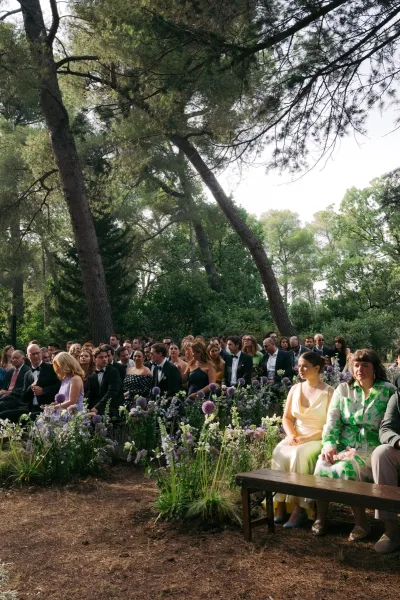Ceremony aisle flowers line a garden ceremony setup, low wildflower clusters flanking wood benches amid pine trees and forest greenery.