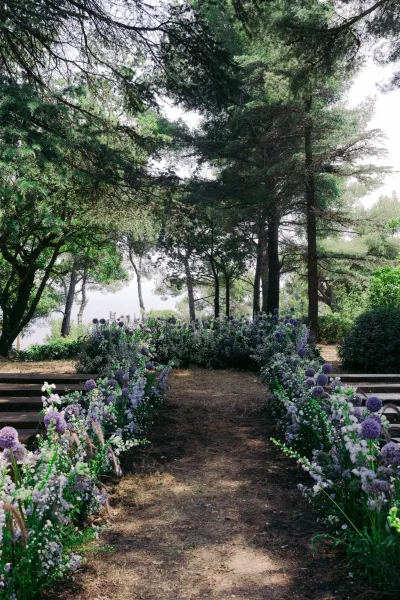 Ceremony aisle design for an outdoor ceremony setup with purple allium blooms edging a dirt path amid forest trees beside calm lake water.