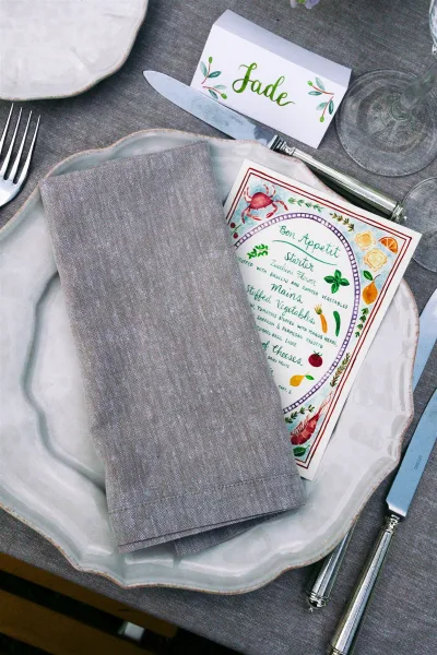 Wedding place setting with a watercolor wedding menu nestled on a scalloped plate, styled simply against a gray tablecloth.