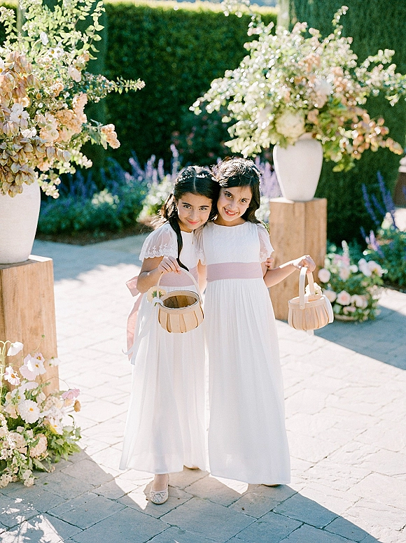 Flower girl portrait of two girls holding wooden flower baskets, in white dresses with pink sashes on a stone patio in a garden setting