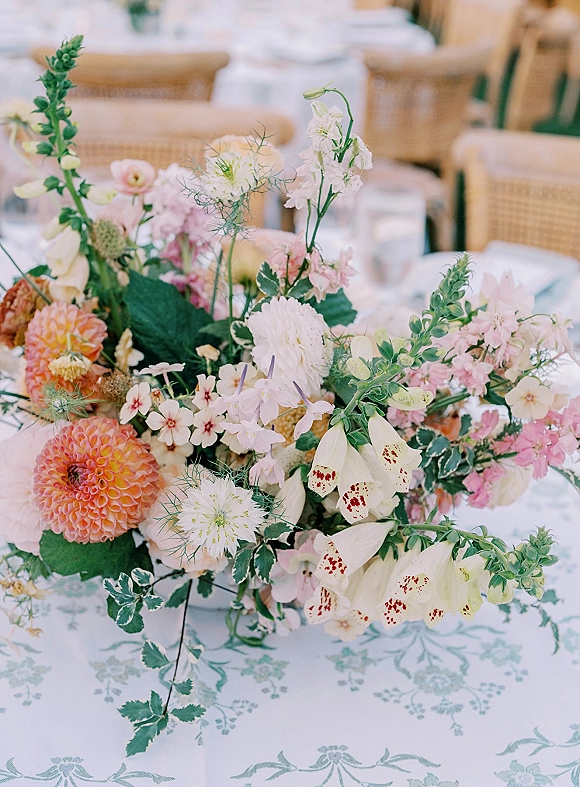 Wedding centerpiece with garden style centerpiece blooms and greenery in a bowl on a patterned tablecloth, with water glasses and reception chairs behind
