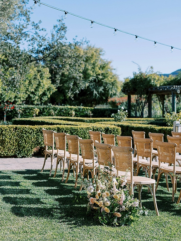 Ceremony setup with outdoor wedding ceremony seating of wood and cane chairs, aisle florals and greenery under string lights on a garden lawn