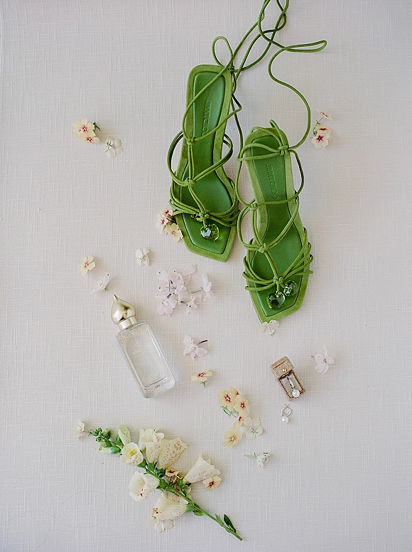 Bridal shoes flat lay with green wedding heels, perfume bottle, drop earrings, ring box, and petals on a white linen backdrop