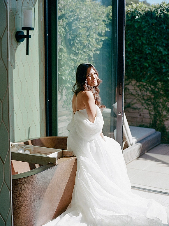 Bridal portrait of a bride in a strapless wedding dress with a tulle skirt, seated by a glass door with patio greenery behind her