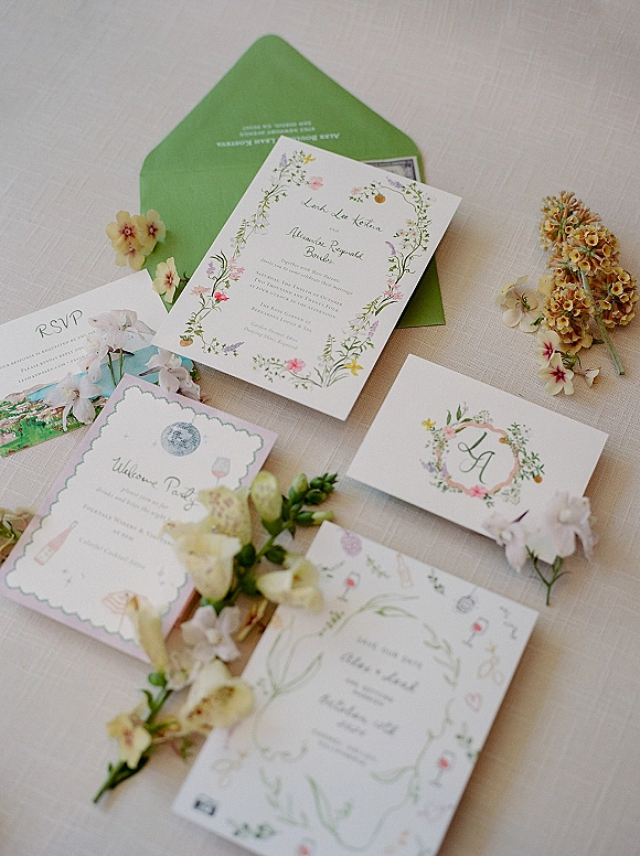 Invitation suite with floral wedding invitations, green envelope, RSVP and details cards, and greenery sprigs arranged on a linen tablecloth