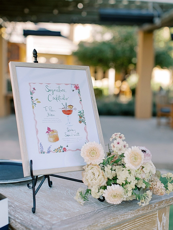 Wedding cocktail sign on an easel with a framed signature cocktails sign, blush floral arrangement, and greenery on an outdoor patio bar table