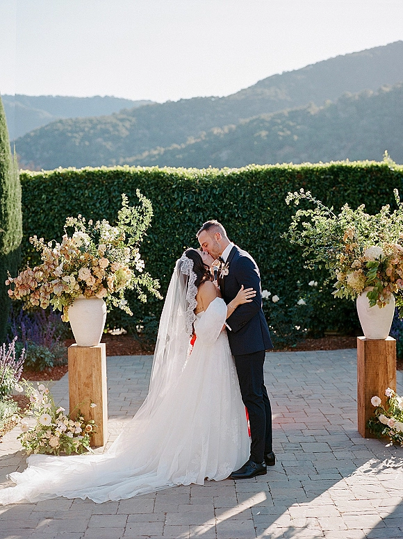 Wedding kiss portrait of bride and groom kissing, her veil blowing beside urn florals on wooden pedestals with mountains behind
