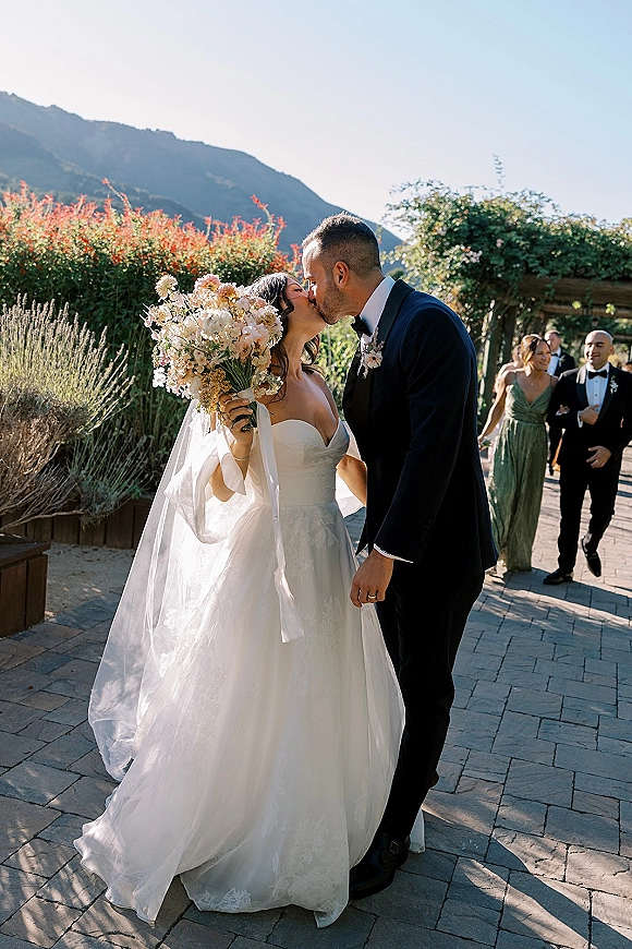 Wedding kiss as bride and groom embrace, her veil flowing and bouquet in hand, on a stone patio with mountain backdrop