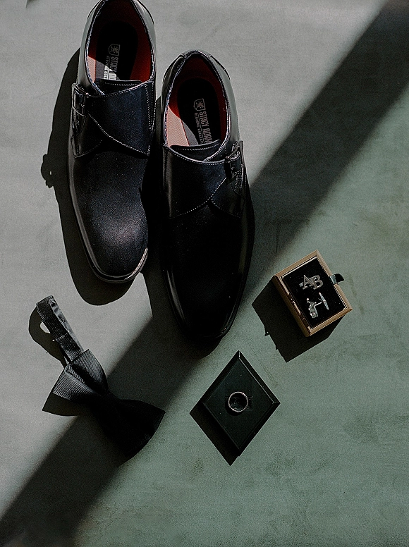 Groom accessories flatlay with black dress shoes, bow tie, cufflinks in box, and wedding ring on a gray floor in natural light shadows