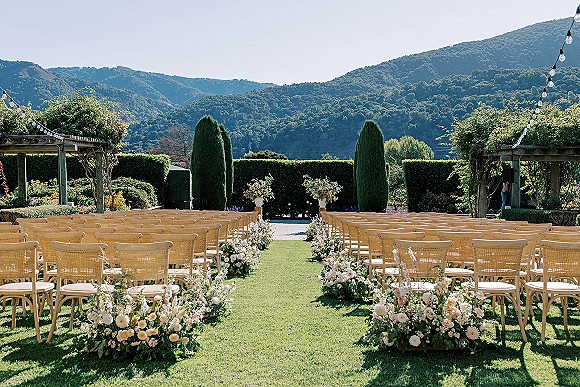 Ceremony setup with outdoor wedding ceremony seating of wood chairs and pastel aisle florals on a lawn, pergola and string lights with mountain view