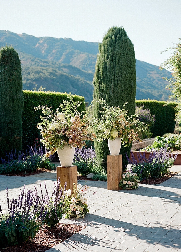Outdoor ceremony decor with wedding altar flowers in white urn vases on wooden pedestals, framed by cypress trees and mountain view backdrop