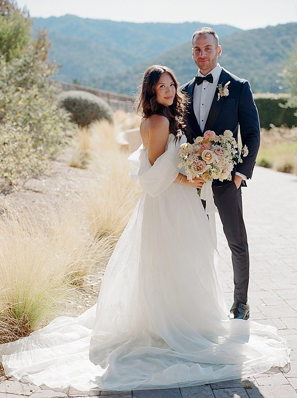 Couple portrait of bride in strapless gown with long train and bow-back holding bouquet beside groom in black tuxedo, mountains behind