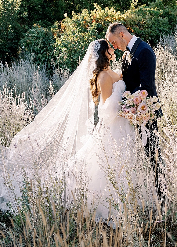 Wedding couple portrait of bride and groom embrace with a lace veil and bouquet, forehead touching in a meadow garden with wildflowers