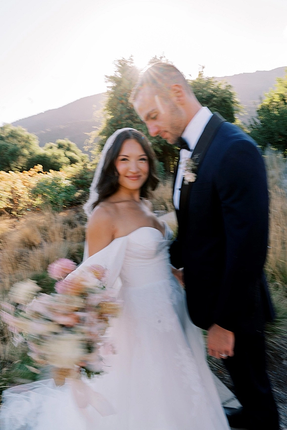 Couple portrait of bride holding bouquet beside groom in tuxedo, her veil catching sun flare against mountains at sunset sky