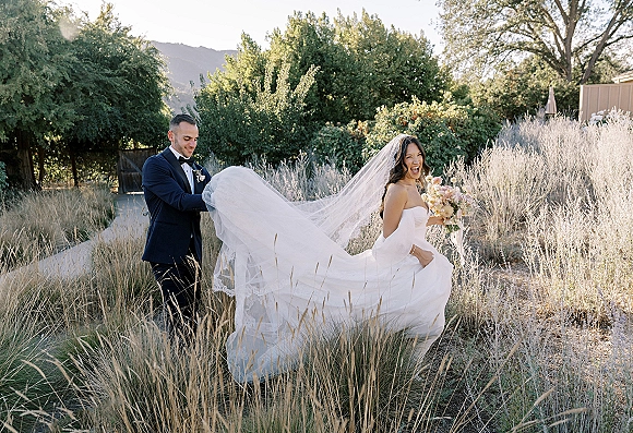 Couple portrait outdoors with bride holding a bouquet and long veil as groom in tuxedo walks beside her on a sunlit garden path
