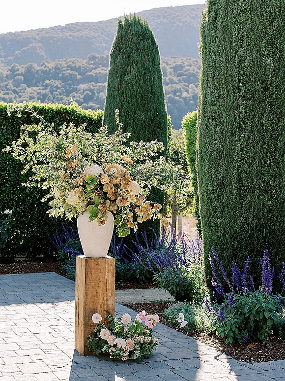 Ceremony floral arrangement in a white urn on a wooden pedestal, with ground blooms beside a stone garden walkway and cypress trees.