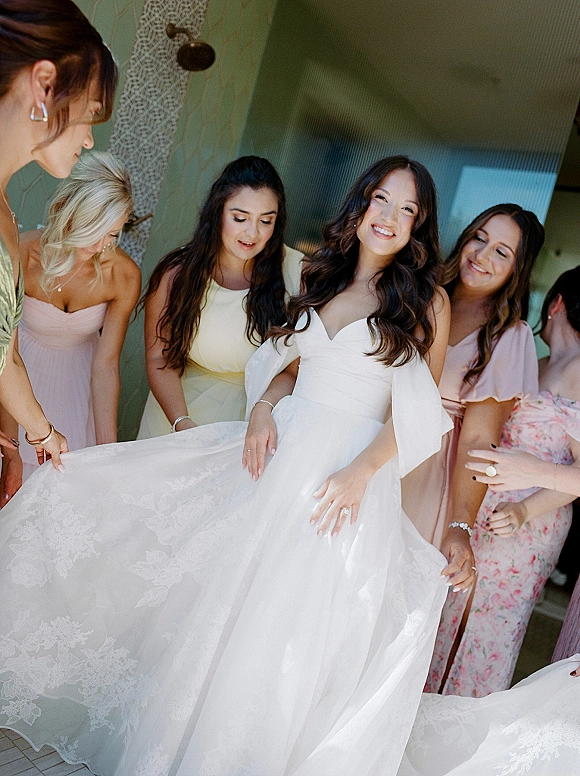 Bride getting ready as bridesmaids help arrange her bridal gown train and off-the-shoulder lace dress in an indoor hallway by a doorway