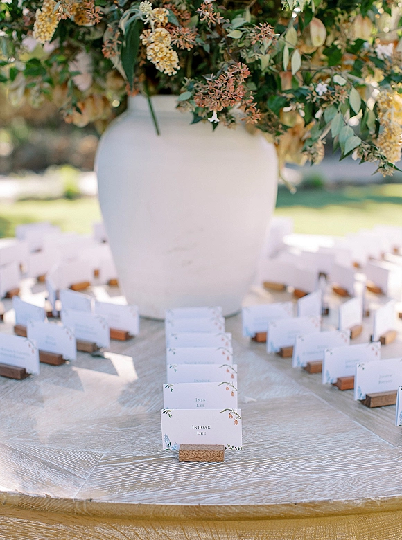 Escort card display with wedding escort cards in wooden holders on a round table, accented by a large white vase of greenery on a sunny lawn