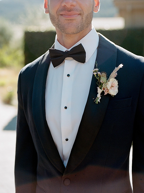 Groom portrait in a black tuxedo with satin shawl lapel and bow tie, wearing a blush boutonniere in sunlit outdoor greenery