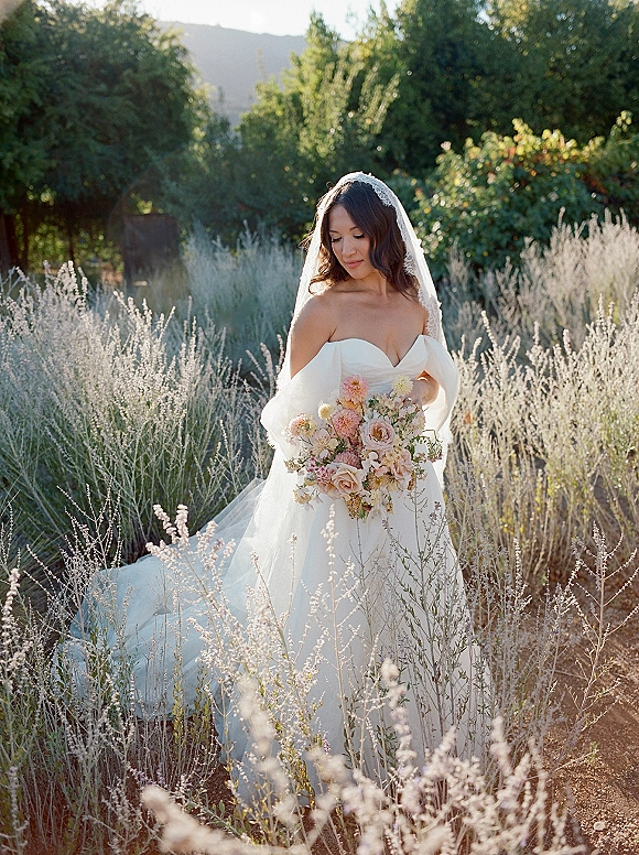 Bridal portrait of a bride holding bouquet, looking down in an off-the-shoulder wedding dress with lace veil amid tall grass and wildflowers