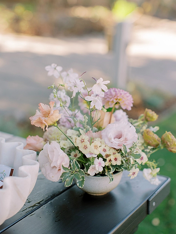 Wedding centerpiece with a pastel wedding centerpiece in a ceramic bowl vase on a ruffled table runner, with escort card and sunlit greenery bokeh behind