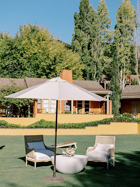 Outdoor lounge setup with wedding lounge seating under a patio umbrella, wicker chairs around a round table with floral centerpiece on a lawn by the house