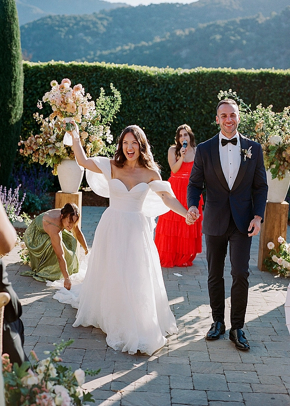 Wedding recessional as bride and groom walk hand in hand, bride lifting bouquet overhead on a paved garden patio with mountain view