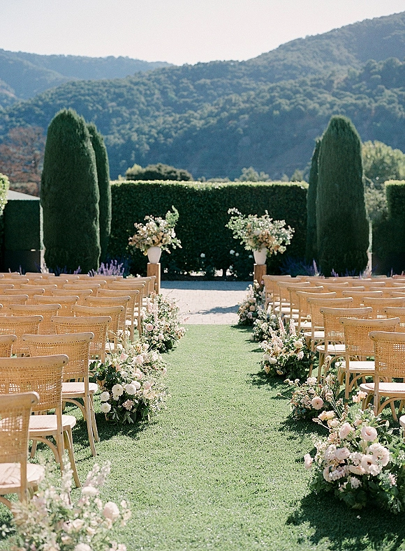 Ceremony setup with outdoor wedding ceremony chairs and floral aisle arrangements on a lawn, framed by hedges, tall trees, and mountain views