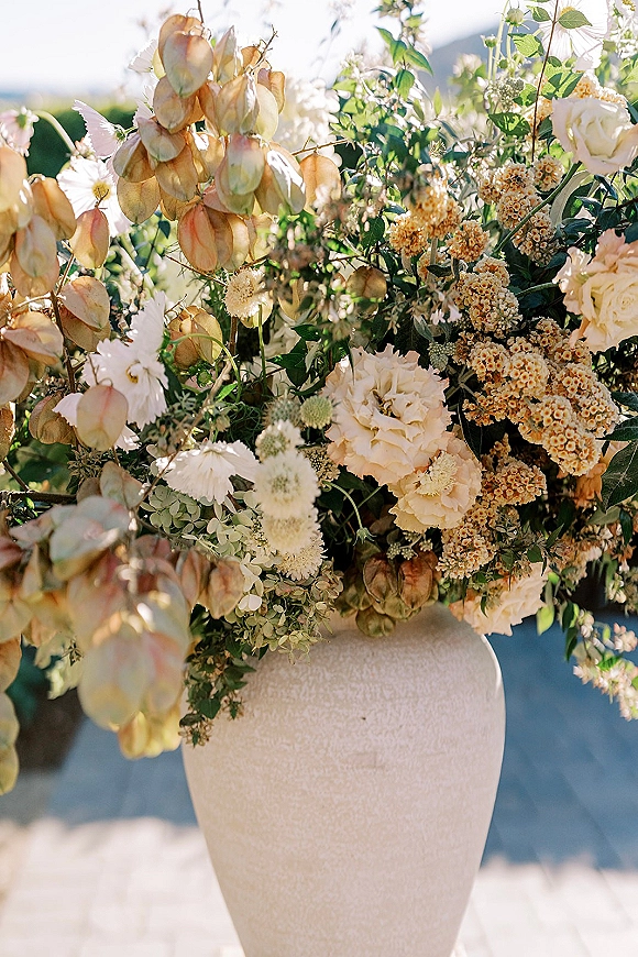 Wedding floral arrangement in a large ceramic urn with white and blush blooms, greenery, baby’s breath and dried pods on a sunlit patio