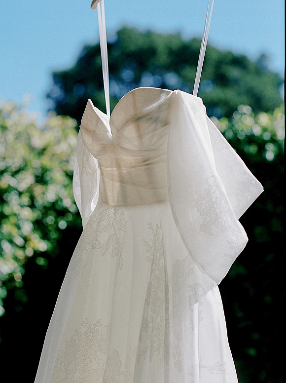 Wedding dress hanging on a hanger, embroidered floral fabric with spaghetti straps and bow detail against garden trees and blue sky