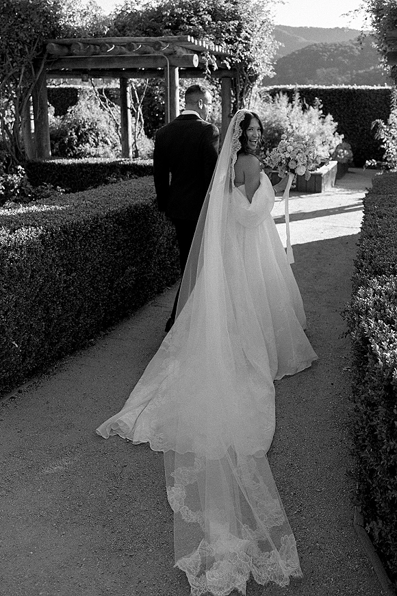 Couple portrait in a black and white wedding portrait as bride looks back holding bouquet, cathedral veil and train on a garden path pergola