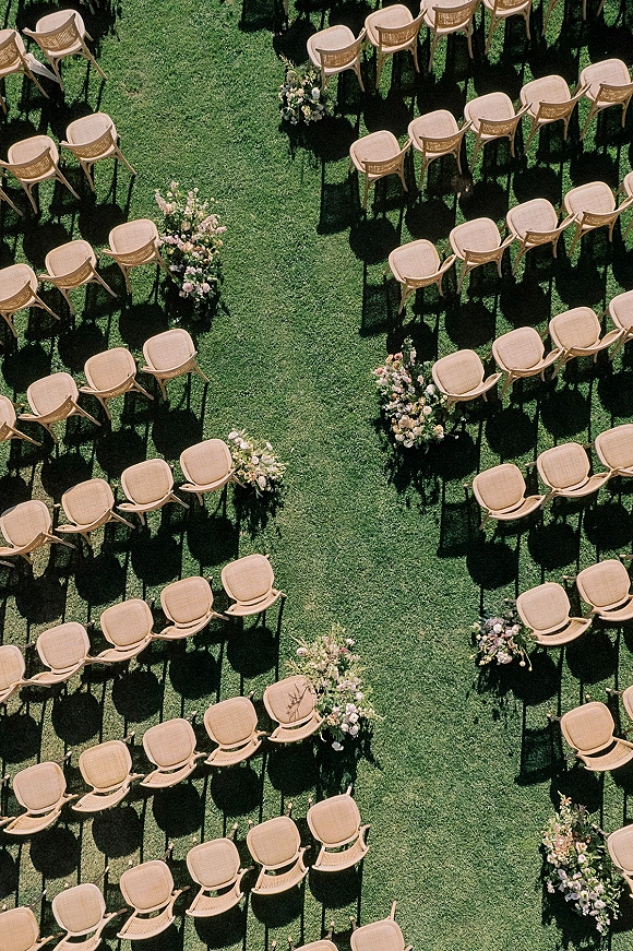 Ceremony seating with wood chairs arranged in rows along a grass lawn aisle, lined with blush and white floral arrangements as markers