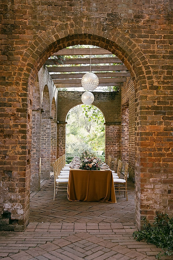 Reception tablescape with a long banquet table setup on a mustard tablecloth, gold Chiavari chairs, florals under lanterns in a brick archway patio