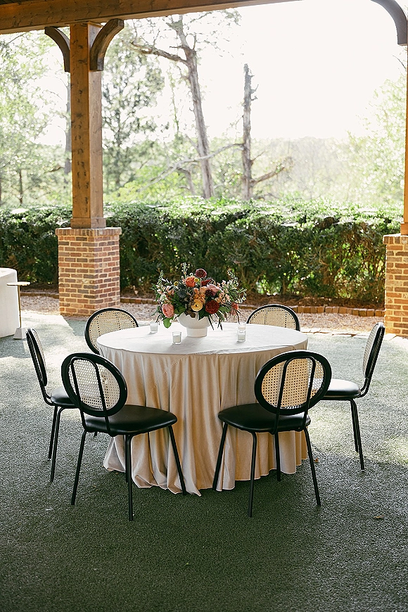Reception tablescape on a round wedding reception table with ivory linen, low floral centerpiece in white vase and votive candles on a covered patio