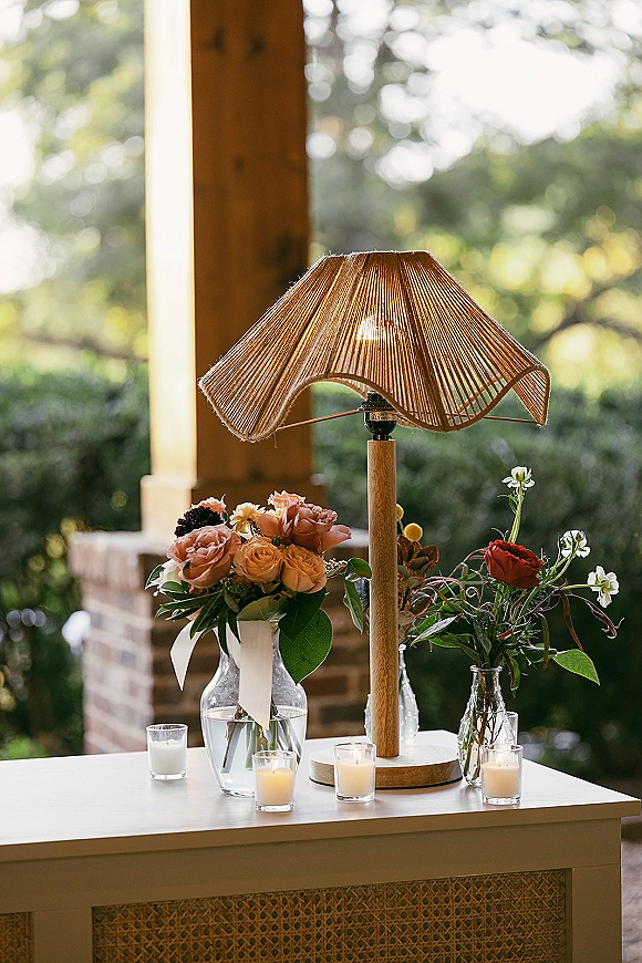Wedding welcome table with table lamp, roses and greenery in glass vases, and votive candles on a porch by brick wall