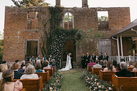 Ceremony moment at an outdoor wedding ceremony as bride and groom kiss beneath a floral arch in ivy-covered brick ruins with wooden pews